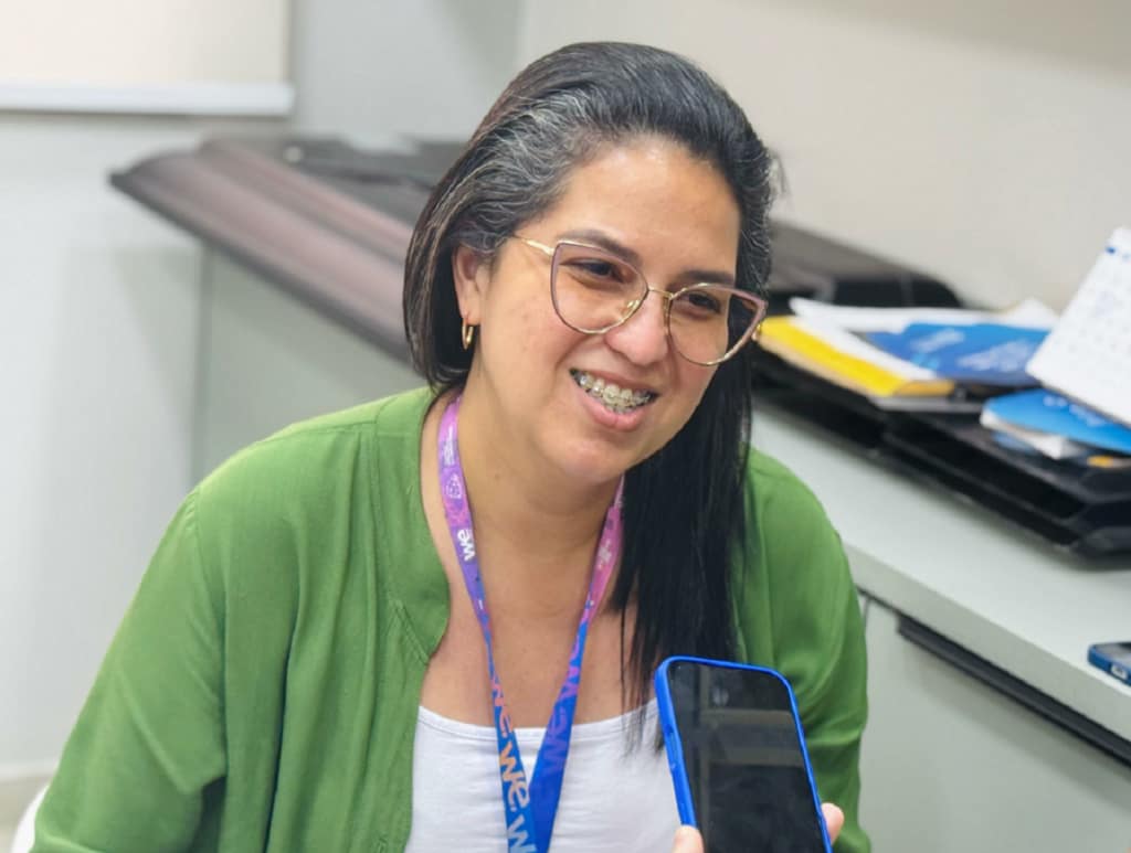 Uma mulher sorrindo. Ela usa um casaco verde e uma blusa branca. Além de estar com os seus óculos e o crachá da empresa Sebrae. Ao fundo, há uma bancada com cadernos e papéis